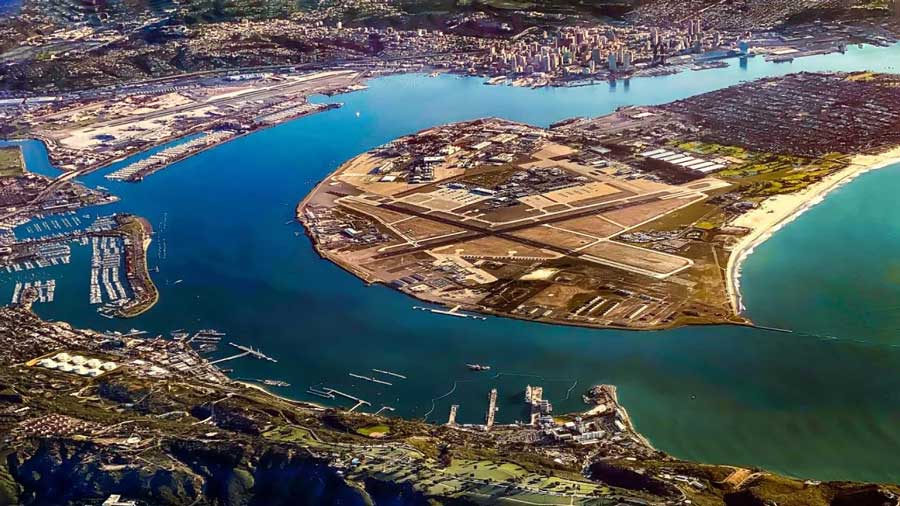 Aerial view of North Island Naval Air Station, located on a peninsula surrounded by water with San Diego’s skyline visible in the background. The airfield, runways, and surrounding military and marina facilities are prominently featured, showcasing the strategic location and layout of the naval base.