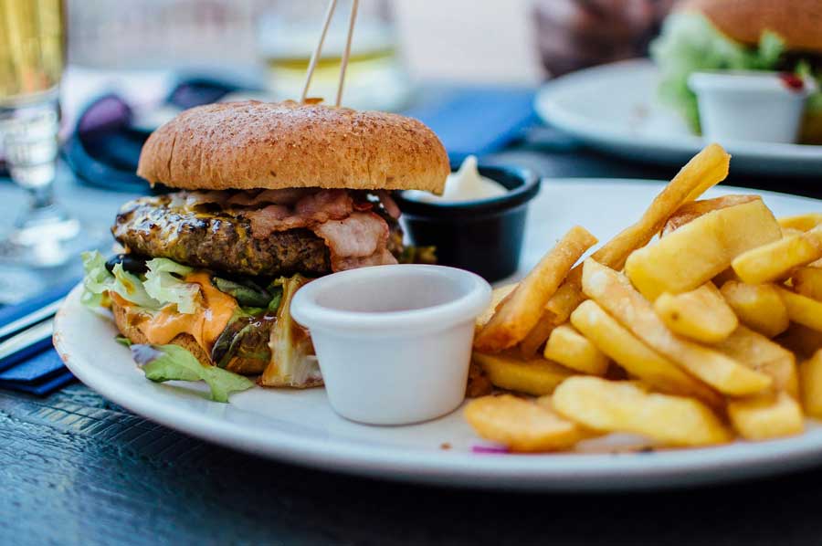 A close-up of a gourmet burger topped with crispy bacon, fresh lettuce, and melted cheese, served with a side of golden fries and dipping sauces. The meal is plated on an outdoor dining table, with another dish and drinks visible in the blurred background, capturing the essence of a delicious food tour experience in San Diego.