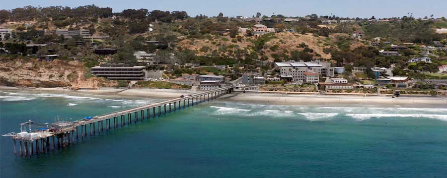 Aerial view of Scripps Institution of Oceanography in La Jolla, California, featuring the long research pier extending into the Pacific Ocean. The coastal campus is surrounded by rolling hills and modern buildings, with waves gently reaching the sandy shore. A hub for marine science and oceanographic research.