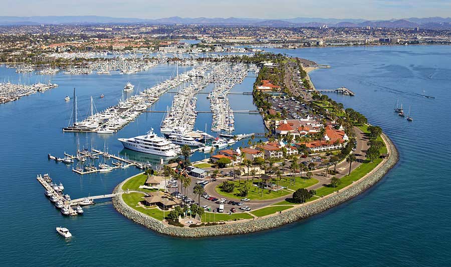 Aerial view of Shelter Island, a scenic waterfront destination with a marina filled with yachts and sailboats. The peninsula features lush greenery, red-roofed buildings, and a curved roadway, with San Diego’s skyline and mountains visible in the background. The vibrant blue waters enhance the picturesque coastal setting.